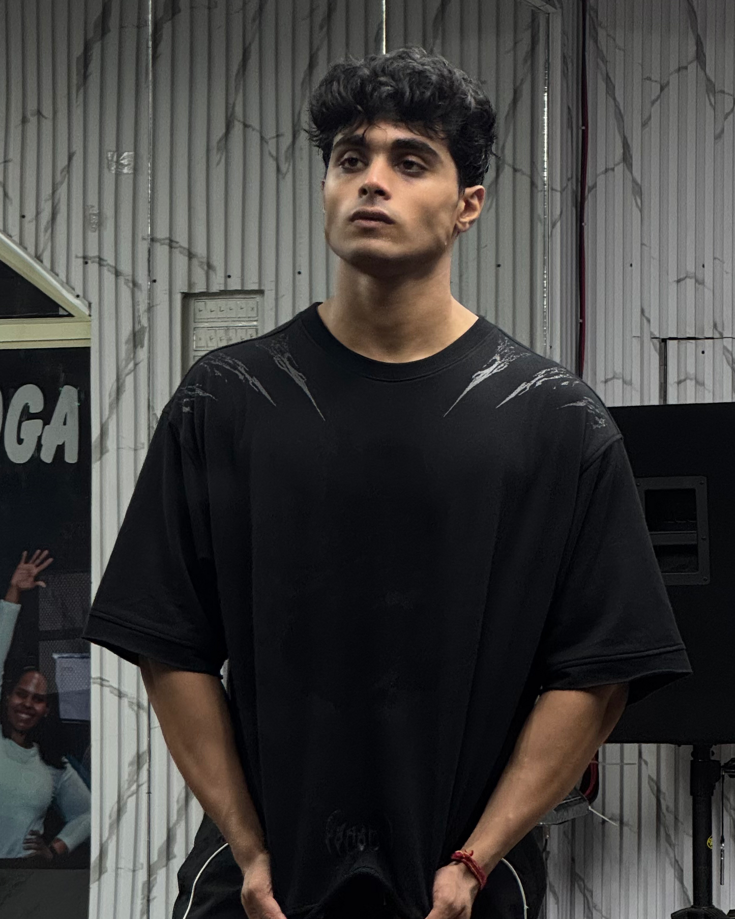 Man wearing a black t-shirt and pants standing in a room with gray walls and a speaker.